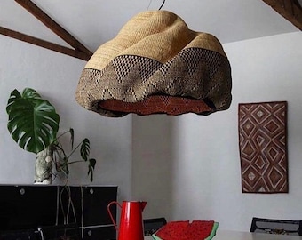 Pode incluir: Uma luminária pendente tecida com um padrão bege e marrom pende do teto. A luz está acima de uma mesa branca com um jarro vermelho e uma fatia de melancia. Uma grande planta verde está ao fundo.