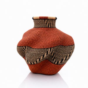 May include: A hand-woven basket with a bulbous, asymmetrical shape and a narrow neck. The exterior features a textured pattern of alternating bands in deep terracotta orange, natural beige, and black, creating a rhythmic, wavy visual effect.