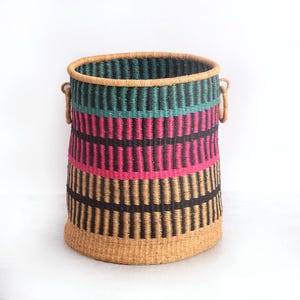 May include: A colourful woven storage basket with a cylindrical shape. The basket has horizontal bands of teal, pink, black, and tan. It has two small handles and a natural-coloured rim. The basket is made of natural fibres.