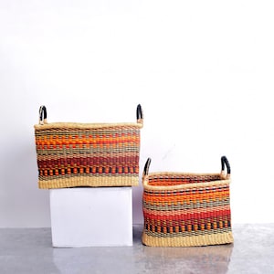 May include: Two woven rectangular baskets with black handles. The baskets feature a vibrant pattern of orange, red, and black horizontal stripes. The baskets are made of natural-colored woven material.