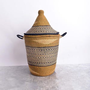 May include: A woven storage basket with a conical lid. The basket is tan with black geometric patterns and a black rim. It has two black handles and is made of natural fibres. The basket is approximately 61 cm tall.