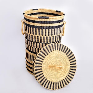 Op de afbeelding: Een hoge, cilindrische geweven mand met een deksel. De mand is gemaakt van natuurlijk bruin en zwart geweven materiaal. Het deksel is rond en heeft een handvat. De mand heeft twee handvatten aan de zijkanten.