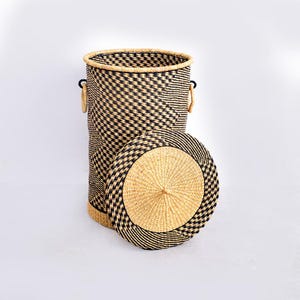 May include: A tall, cylindrical laundry basket with a matching lid. The basket is woven with a black and natural fibre pattern. The lid is round with a central knob. The basket has two handles.