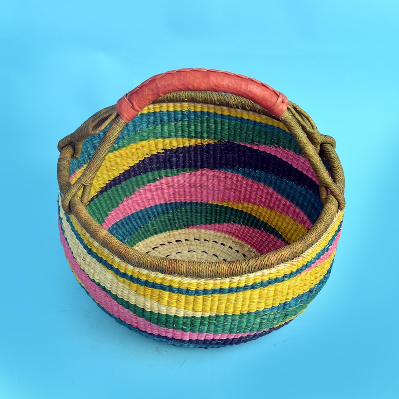 Peut inclure: Un panier rond tiss&eacute; avec un motif ray&eacute; color&eacute; dans les tons de rose, jaune, vert et bleu. Le panier a un bord marron et une poign&eacute;e en cuir rouge. Le panier est pos&eacute; sur un fond bleu clair.