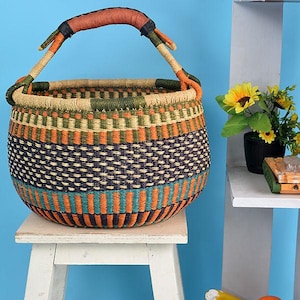 Peut inclure: Un panier rond tissé avec une anse. Le panier présente un motif coloré de rayures horizontales orange, turquoise, vertes et bleu marine. La poignée est enveloppée de cuir marron. Le panier est posé sur un petit tabouret blanc.