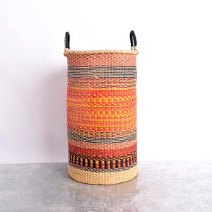 Può includere: Un cesto alto e cilindrico intrecciato con una vivace combinazione di colori. Il cesto presenta fasce orizzontali rosse, arancioni, gialle e nere. Ha due manici neri ed è realizzato con fibre naturali. Il cesto è alto circa 76 cm.