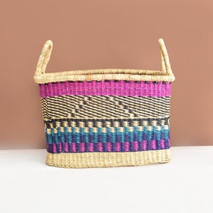 May include: A rectangular woven basket with two handles. The basket features a vibrant design with bands of pink, black, teal, and blue. The base and handles are a natural beige color.
