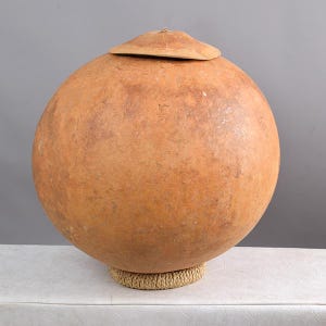 May include: A large, round, brown gourd with a lid. The gourd is sitting on a woven base.