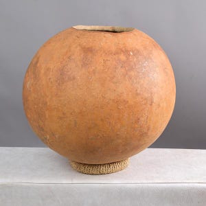 May include: A large, round, brown gourd with a natural, rough texture. The gourd is sitting on a woven base.