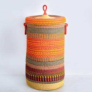 May include: A tall, cylindrical woven basket with a lid. The basket features a vibrant color scheme of red, orange, yellow, black, and brown horizontal stripes. It has two red handles and a red loop on the lid. The basket is approximately 30 inches tall.