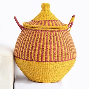 May include: A yellow and burgundy woven storage basket with a lid. The basket has a rounded shape with a lid and two handles. The basket is made of natural materials. The basket is sitting on a white surface. The basket is made of natural materials.