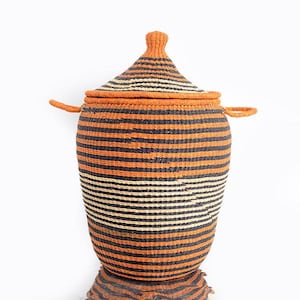 May include: A handwoven storage basket with a lid. The basket has a cylindrical body and a conical lid, with two small handles. It features horizontal stripes in orange, black, and cream. The basket is made of natural fibres.
