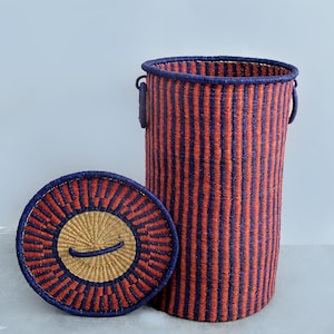 May include: A tall, cylindrical woven basket with a matching lid. The basket and lid feature vertical orange and blue stripes. The lid has a circular design with a tan centre and a blue border. The basket has two handles.