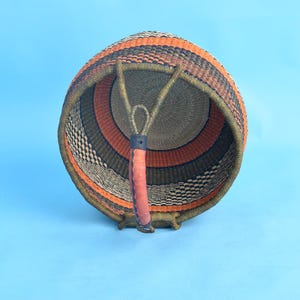 May include: A round, woven basket with a handle. The basket features a color scheme of orange, black, and olive green. The handle is made of leather and is attached to the basket with a loop. The basket is likely used for storage or decoration.