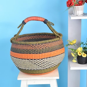 May include: A round, handwoven basket with a sturdy handle. The basket features horizontal stripes in orange, olive green, and black. The handle is wrapped in orange leather. The basket is sitting on a white stool.