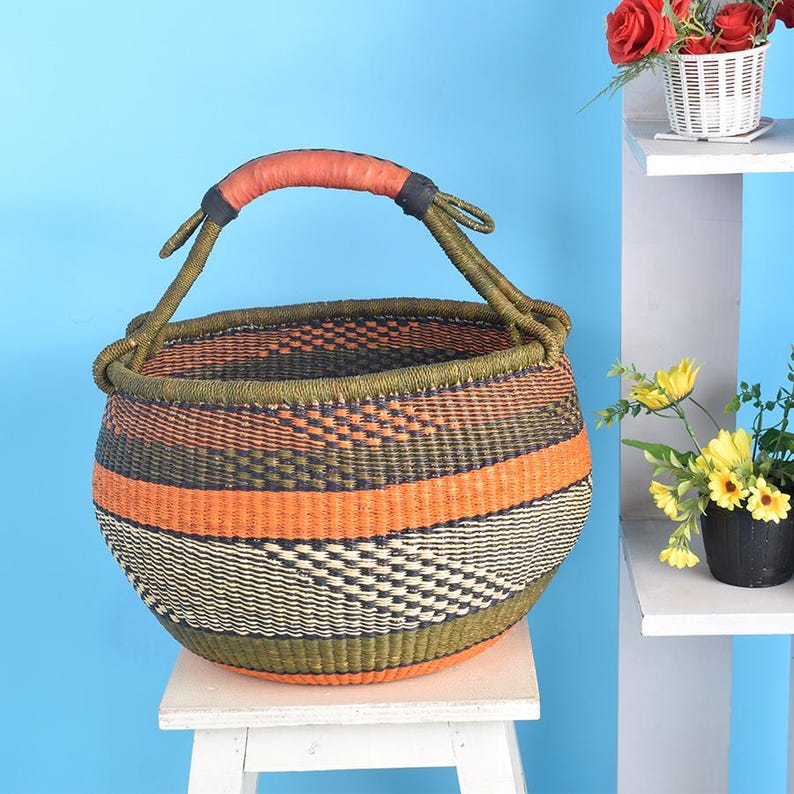 May include: A round, handwoven basket with a sturdy handle. The basket features horizontal stripes in orange, olive green, and black. The handle is wrapped in orange leather. The basket is sitting on a white stool.