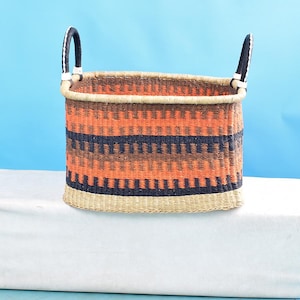 May include: Woven storage basket with a rectangular shape and two arched handles. The basket features a pattern of orange, brown, and navy blue horizontal stripes. The rim and base are a natural beige color.