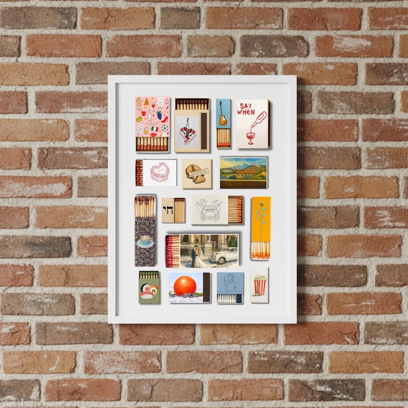 May include: A framed collection of matchbox art, featuring various designs and illustrations. The artwork includes images of a guitar, a wine bottle, a landscape, and the text "SAY WHEN". The frame is white, and the background is a brick wall.