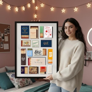 May include: A framed collection of vintage matchboxes, featuring various designs and brands. The matchboxes are arranged in a grid, showcasing diverse colors, fonts, and graphics. Some boxes display text like "ROLOV" and "E-ET'S". The frame is held by a person.
