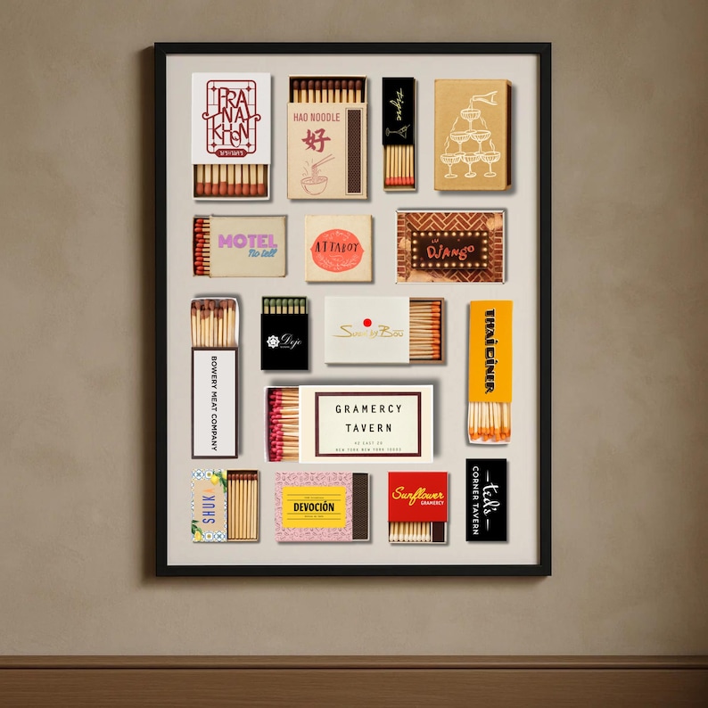 May include: A framed print featuring a collection of vintage matchboxes. The matchboxes display various designs and text, including "PRA NA KHEN", "HAO NOODLE", "MOTEL", "ATTALOT", "DJANGO", "BOWERY MEAT COMPANY", "SHUK", "GRAMERCY TAVERN", "THAI DINER", "DEVOCI&Oacute;N", and "Sunflower".