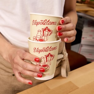 May include: Two cream-colored paper coffee cups stacked together, each with the word "Napoletana" in red script. Red illustrations of a coffee pot, milk bottle, and coffee cup are also visible. The cups are held by a person with red nail polish.