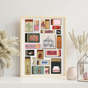 May include: A framed artwork showcasing diverse matchbox designs. The designs include a cactus, a mushroom, a suitcase, a gazebo, and the text "Cheers" and "Be Kind". The artwork is displayed on a white surface.