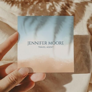 May include: Square business card with a watercolour beach scene. The top is light blue, transitioning to beige at the bottom. The card features the text "JENNIFER MOORE" and "TRAVEL AGENT" in a dark blue font.