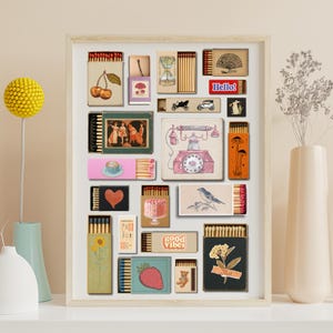 May include: A framed artwork showcasing a collection of matchboxes with retro illustrations. The matchboxes display cherries, a mushroom, an hourglass, a vintage car, a fan, a rabbit, a couple dancing, a teacup, a heart, a cake, a bird, a sunflower, a strawberry, a teddy bear, and the words "Hello!" and "good vibes."