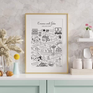 May include: A framed black and white illustration titled "Emma and Jake OUR LOVE STORY." The artwork depicts a timeline of events with various icons, including a movie camera, mountains, and a boat, all within a gold frame.