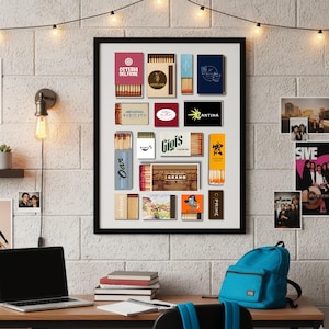 May include: A framed collection of matchboxes with various designs and logos. The matchboxes are arranged in a grid, showcasing different brands and styles. The frame is black, and the background is a white brick wall. A desk with a laptop, books, and a backpack is in the foreground.