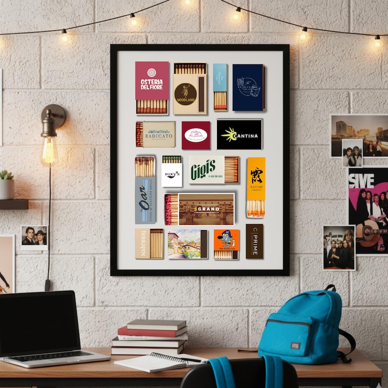 May include: A framed collection of matchboxes with various designs and logos. The matchboxes are arranged in a grid, showcasing different brands and styles. The frame is black, and the background is a white brick wall. A desk with a laptop, books, and a backpack is in the foreground.