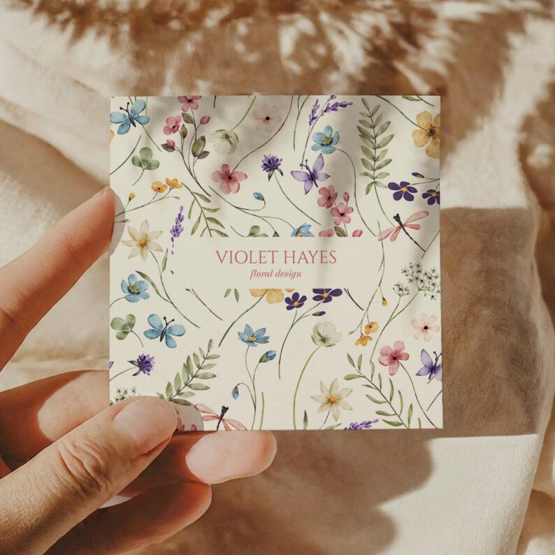 May include: A square business card with a cream background, adorned with a floral pattern in pink, blue, purple, and yellow. The card features the text "VIOLET HAYES floral design" within a rectangular banner.