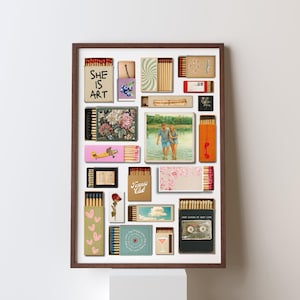 May include: A framed artwork composed of various matchboxes. The matchboxes display diverse designs, including floral patterns, a photograph of a couple, and the text "SHE IS ART". The artwork is set against a white backdrop.
