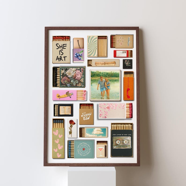May include: A framed artwork composed of various matchboxes. The matchboxes display diverse designs, including floral patterns, a photograph of a couple, and the text "SHE IS ART". The artwork is set against a white backdrop.