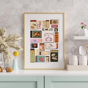 May include: A framed art print displaying a collage of vintage matchbox designs. The matchboxes feature diverse illustrations, such as cherries, a teacup, a bird, and the text "Hello!". The artwork is framed in gold and displayed on a white surface.