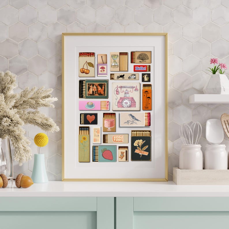 May include: A framed art print displaying a collage of vintage matchbox designs. The matchboxes feature diverse illustrations, such as cherries, a teacup, a bird, and the text "Hello!". The artwork is framed in gold and displayed on a white surface.