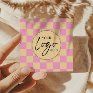 May include: Square business card featuring a pink and beige chequered design. A circular logo area in the centre displays the text "YOUR Logo HERE" in a handwritten font. The card is held in a person's hand.