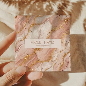 May include: A square business card with a marble design in shades of pink and gold. The card features the text "VIOLET HAYES wedding planner" in a light brown rectangle.