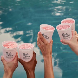 May include: Several white cups filled with pink slushies are held up. The cups have the words "Bride Squad" printed on them in black. The background is a blue swimming pool.