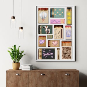 May include: A framed artwork showcasing diverse matchbox designs. The designs include a hot air balloon, cow, floral patterns, and the word "Gratitude". The artwork is displayed on a wooden cabinet with a plant and hanging lights.