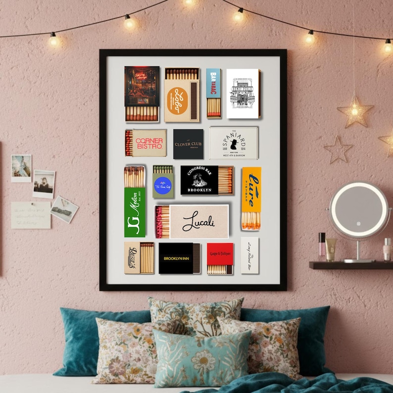 May include: A framed collection of vintage matchboxes, featuring various designs and logos. The matchboxes are arranged on a pink wall, with string lights and decorative pillows in the foreground. The frame is black.