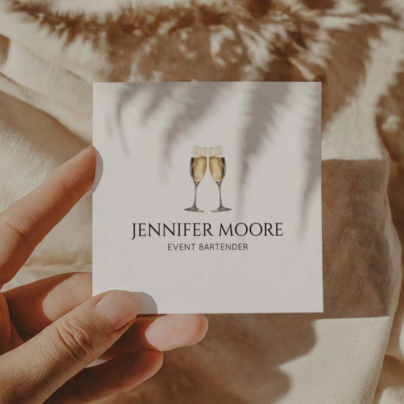 May include: A white business card with the name "JENNIFER MOORE" and the title "EVENT BARTENDER" in black text. Two champagne flutes with a bubbly liquid are illustrated above the name. The card is held by a hand, with a shadow overlay.