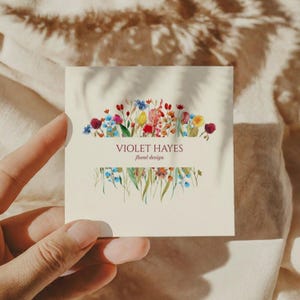 May include: A square business card with the text "VIOLET HAYES floral design" and a watercolour floral design. The card is held by a hand and features a cream background with colourful flowers.
