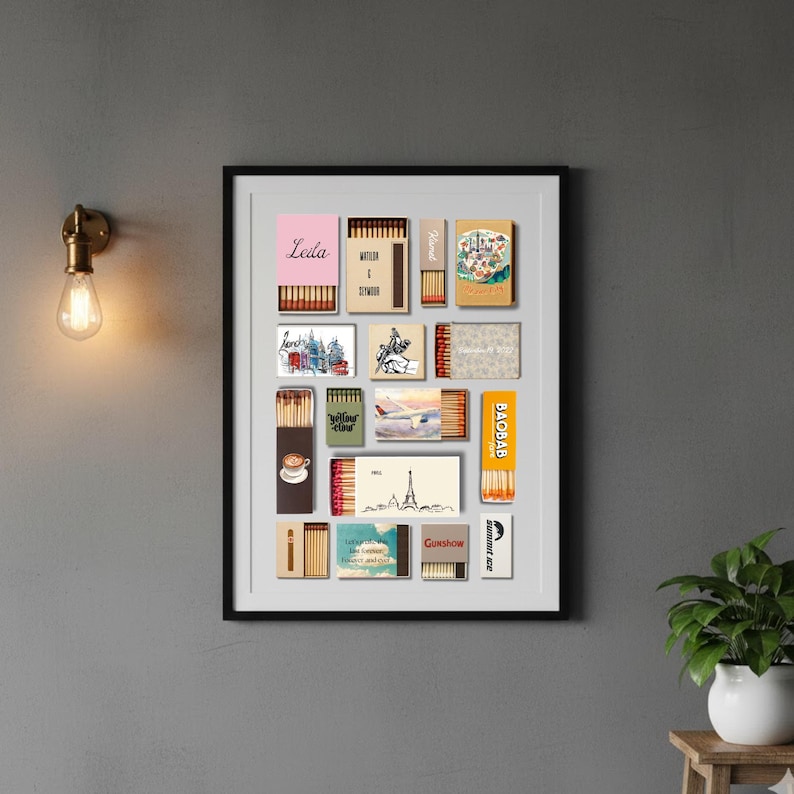 May include: A framed print featuring a collection of vintage matchboxes. The matchboxes display various designs, including travel scenes, text, and illustrations. The frame is black, and the artwork is mounted on a gray wall.