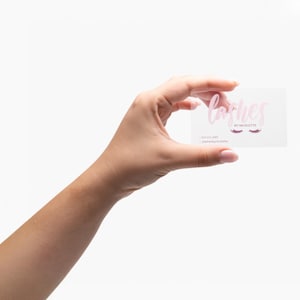 May include: A clear business card held in a hand, featuring the word "lashes" in pink script, with the text "by Nicolette" below. Two pink eyelash designs are also visible. Contact information is printed below.