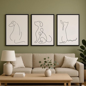 May include: Three framed prints with minimalist dog line drawings. Each black-framed print shows a different dog pose in a continuous black line on a cream background. The artwork is displayed above a beige sofa, creating a modern aesthetic.