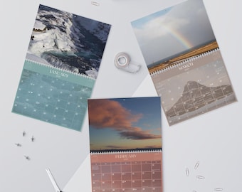 May include: Three wall calendars with different nature scenes. The January calendar features a waterfall in Iceland, the February calendar features a sunset with a large cloud, and the March calendar features a rainbow over a beach.
