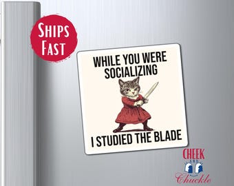 May include: A white refrigerator magnet with a black and white illustration of a cat holding a sword. The text on the magnet reads "While you were socializing I studied the blade".