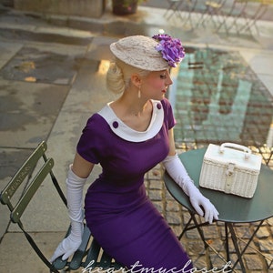 May include: A woman wearing a purple dress and a white hat with purple flowers sits on a chair with a white wicker basket on a table. The table is made of metal and has a green top. The woman is wearing white gloves and black and white shoes. The background is a cobblestone street.