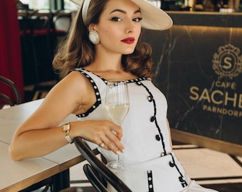 May include: A woman wearing a white dress with black polka dots and a white hat sits at a table in a cafe. She is holding a glass of champagne. The cafe is called "Cafe Sache Parndorf".